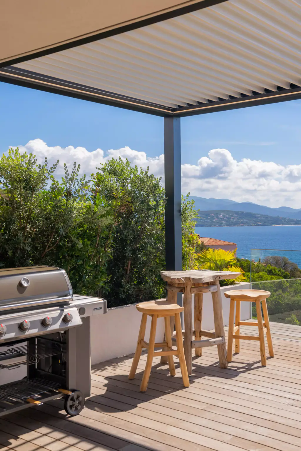 Sous l’angle de la pergola : lames claires, barbecue, mange-debout et tabourets en teck, le golfe au loin.