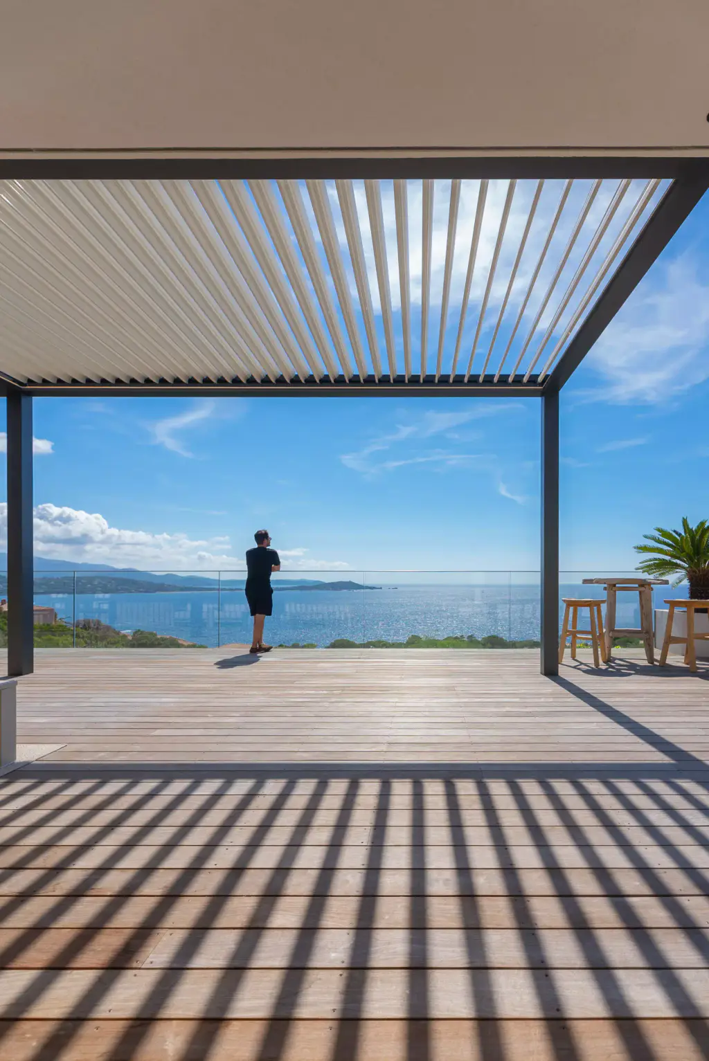 Sous une pergola bioclimatique aux lames entrouvertes, un homme de dos regarde le golfe depuis le garde-corps vitré d’une terrasse en bois rayée d’ombres.