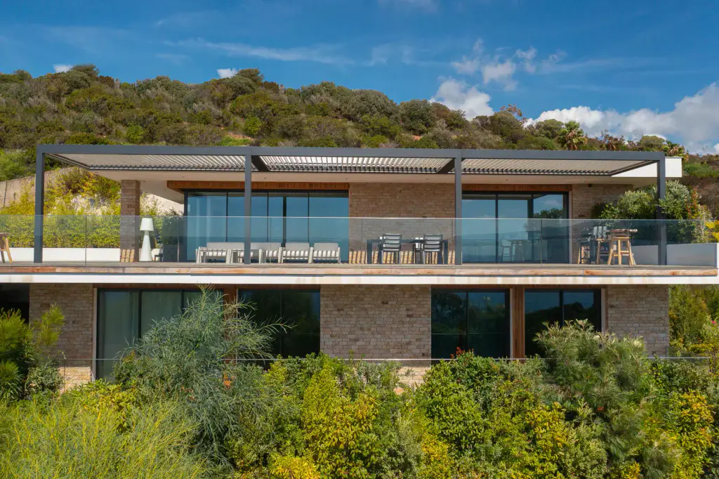 Bioclimatic pergola with an anthracite frame seen head on by drone on the upper terrace of a stone villa: white lounge, table, glass balustrade, maquis in the foreground.