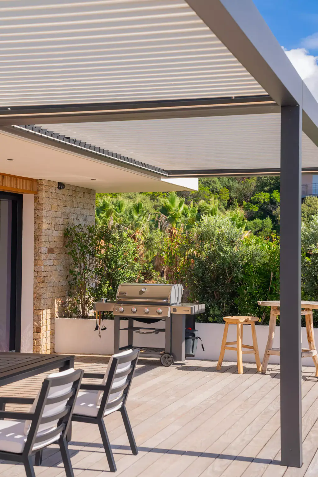 Corner of the pergola (anthracite posts, light-coloured louvres closed) above the barbecue area and teak stools; stone wall, dense greenery, two aluminium chairs in the foreground.