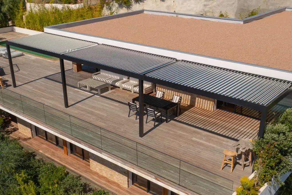 Pergola bioclimatique vue par drone, lames fermées : la toiture couvre la table anthracite et le salon blanc sur le deck en bois, garde-corps en verre, toit gravillonné.