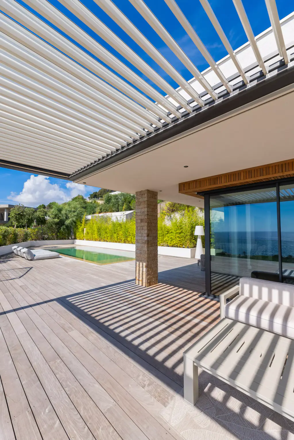 Lames orientées ouvertes vues de dessous contre un ciel bleu profond, ombres rayées sur le deck, baie vitrée et piscine au fond.