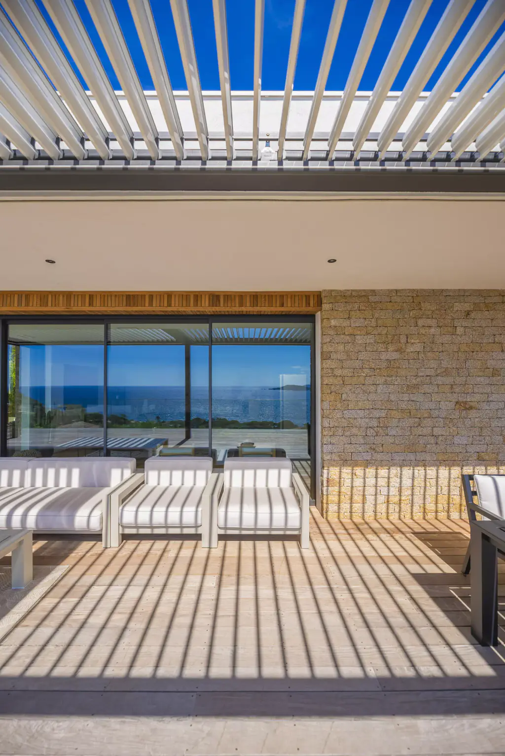 Open louvres at the top of the frame, glazed façade reflecting the sea, light sofas, floor striped with shadow.