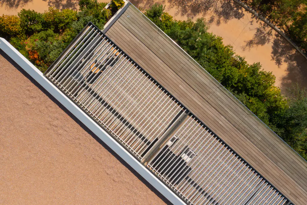 Light-coloured louvres of the bioclimatic pergola open, seen from directly above by drone: the table and lounge show through, wooden deck, gravel roof, maquis.
