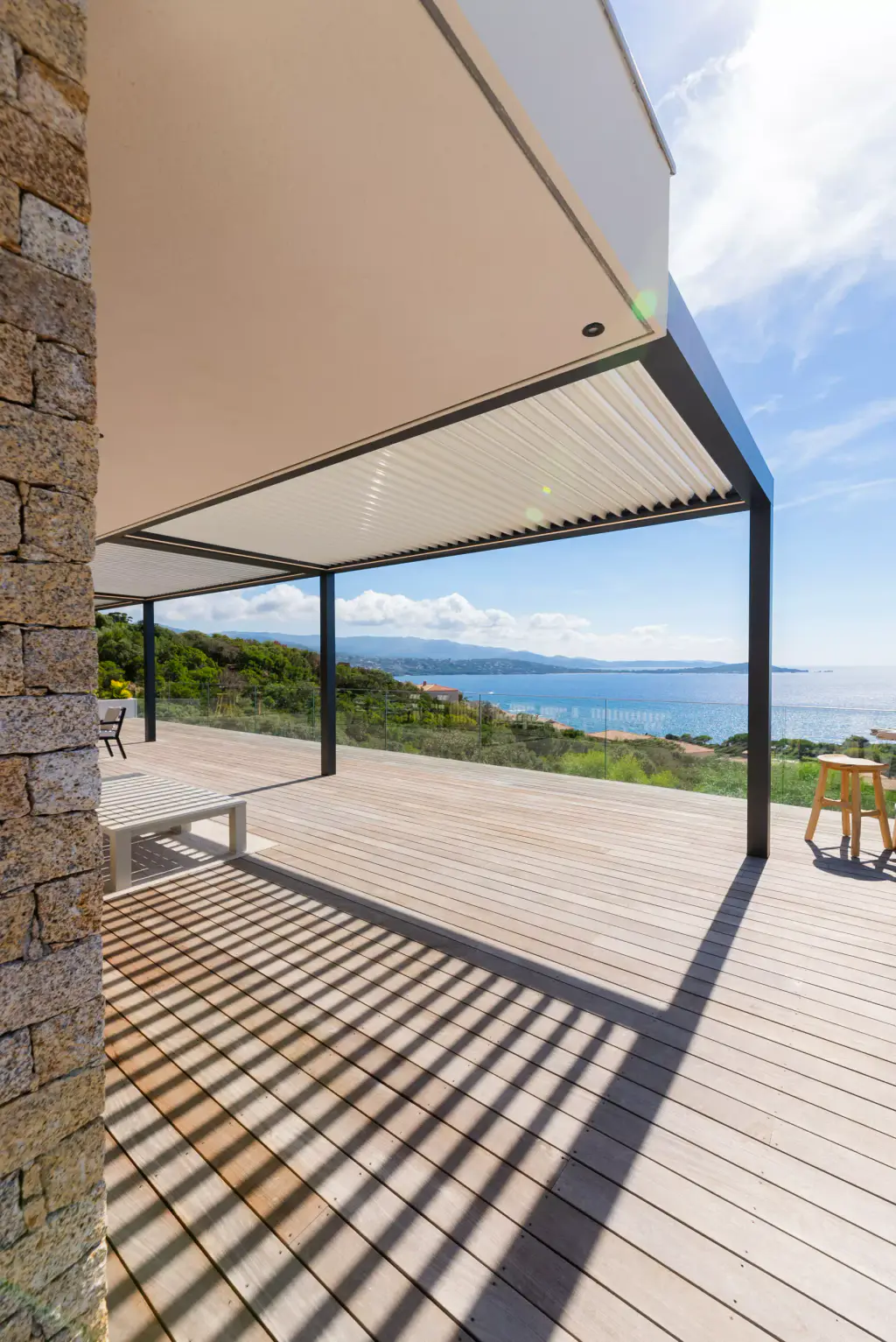 Stone pillar framing the left, pergola in receding perspective, louvre shadows on the deck, sea horizon.