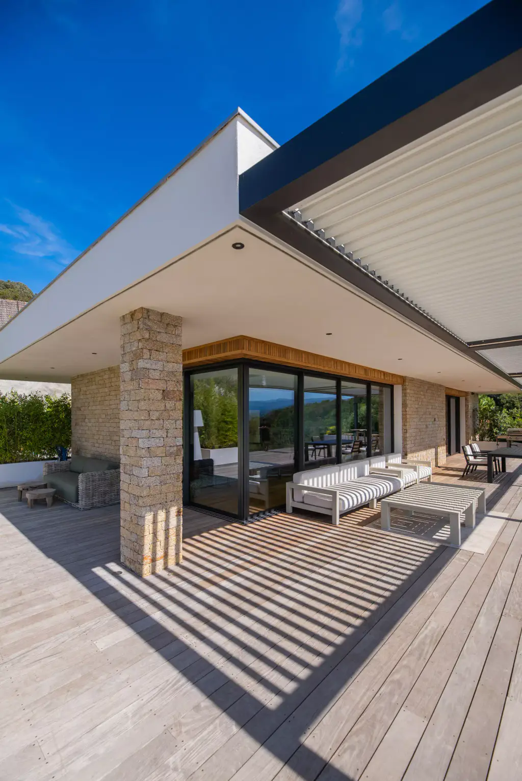 Low-angle view of the corner of the villa: stone pillar, pergola, outdoor lounge with shadow-striped cushions.