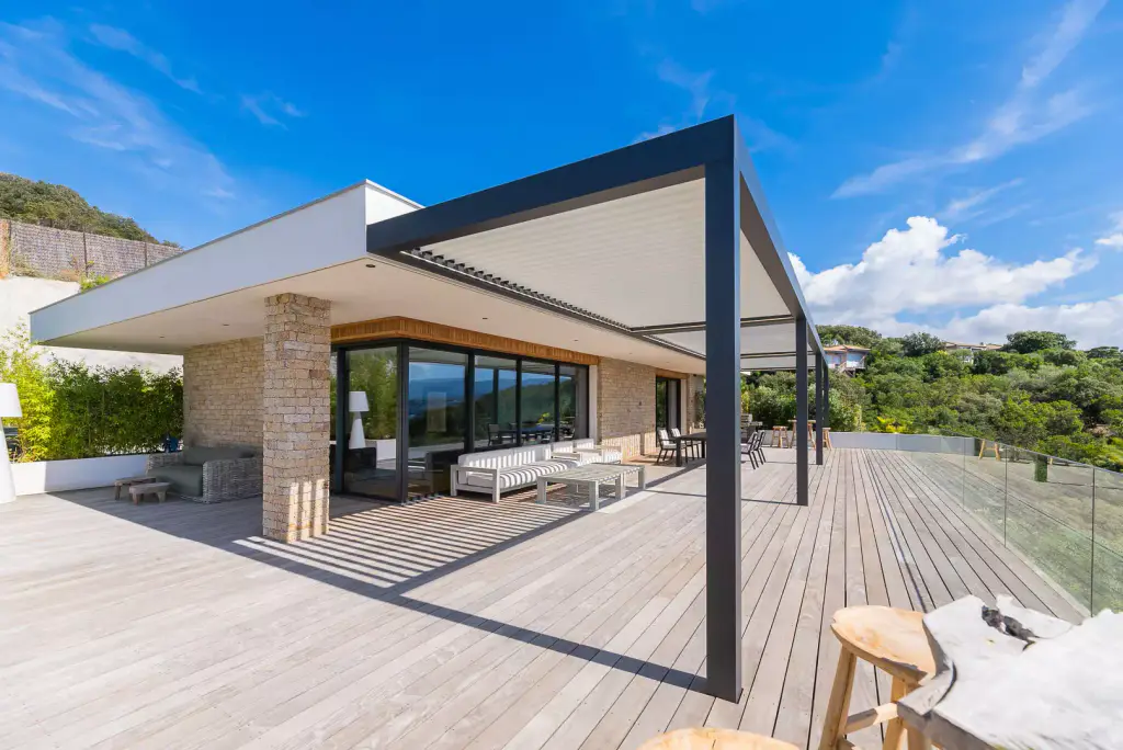 Pergola bioclimatique à cadre anthracite en porte-à-faux à l’angle de la villa, grand deck en bois, garde-corps en verre, baies vitrées et maquis sous un ciel bleu.