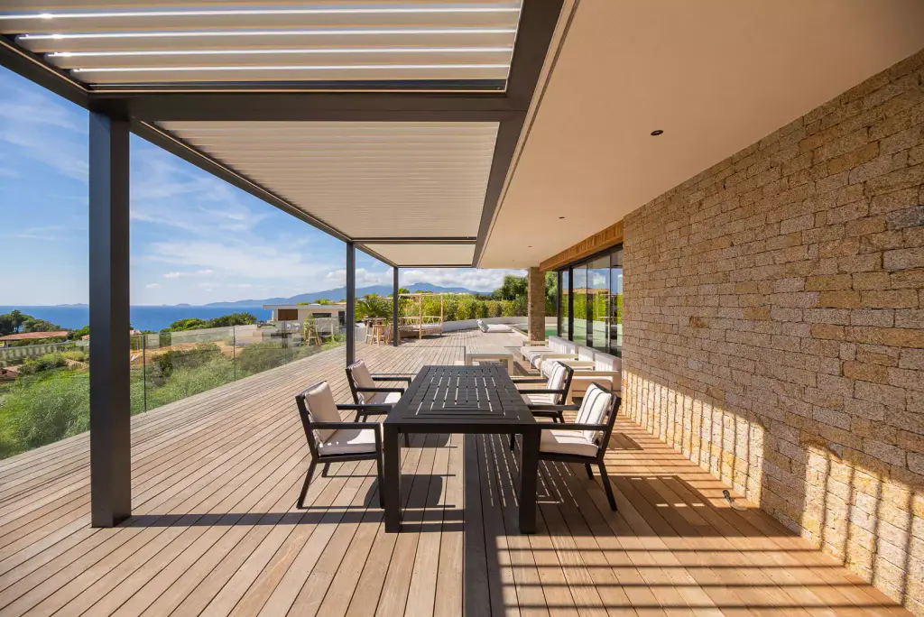 Long perspective under the pergola: table, dry-stone wall on the right, striped shadows, sea on the left.