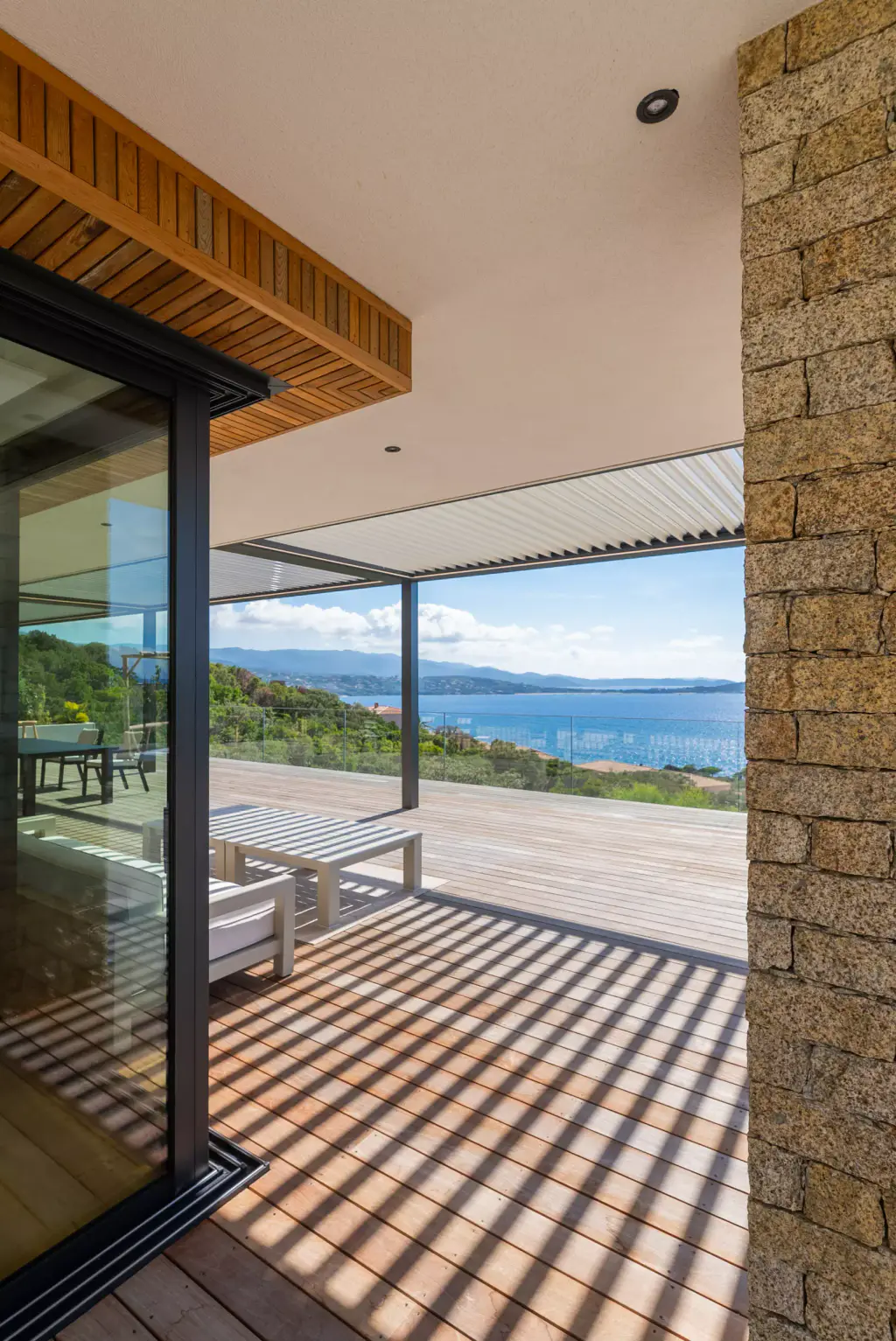 View from the glazing: timber-clad soffit, stone pillar, pergola and sea in the background, striped shadows on the floor.