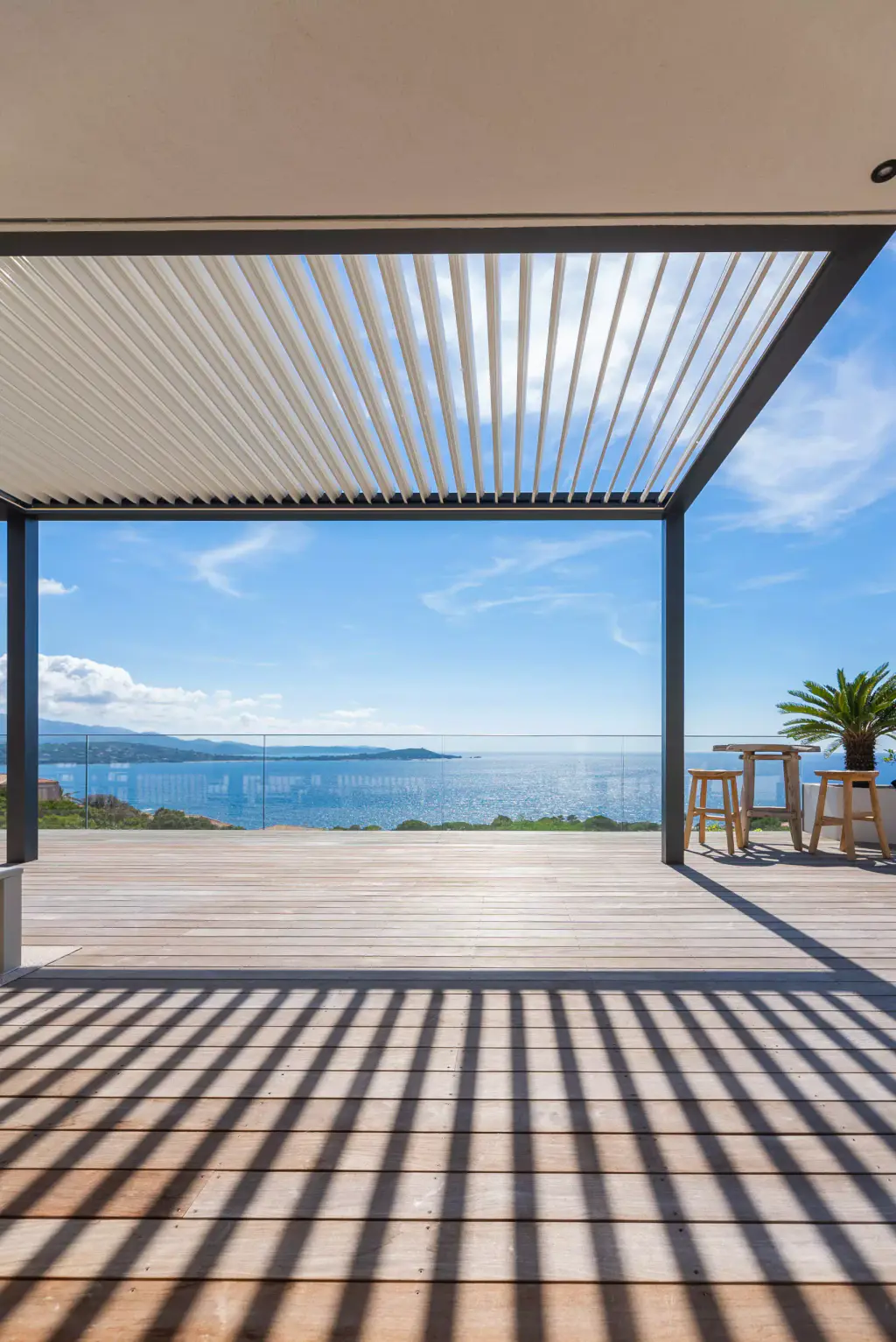 Symmetrical front framing: half-open louvres, anthracite frame, gulf, deck striped with shadow.