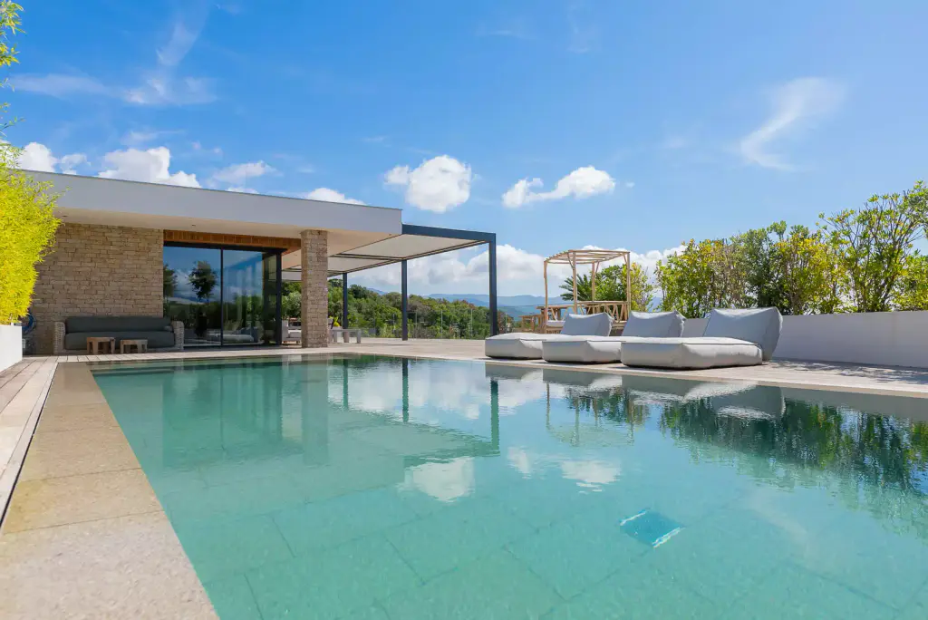 Piscine à l’eau turquoise au premier plan, plage en pierre claire, bains de soleil blancs et lit à baldaquin, villa contemporaine en pierre et pergola au fond.
