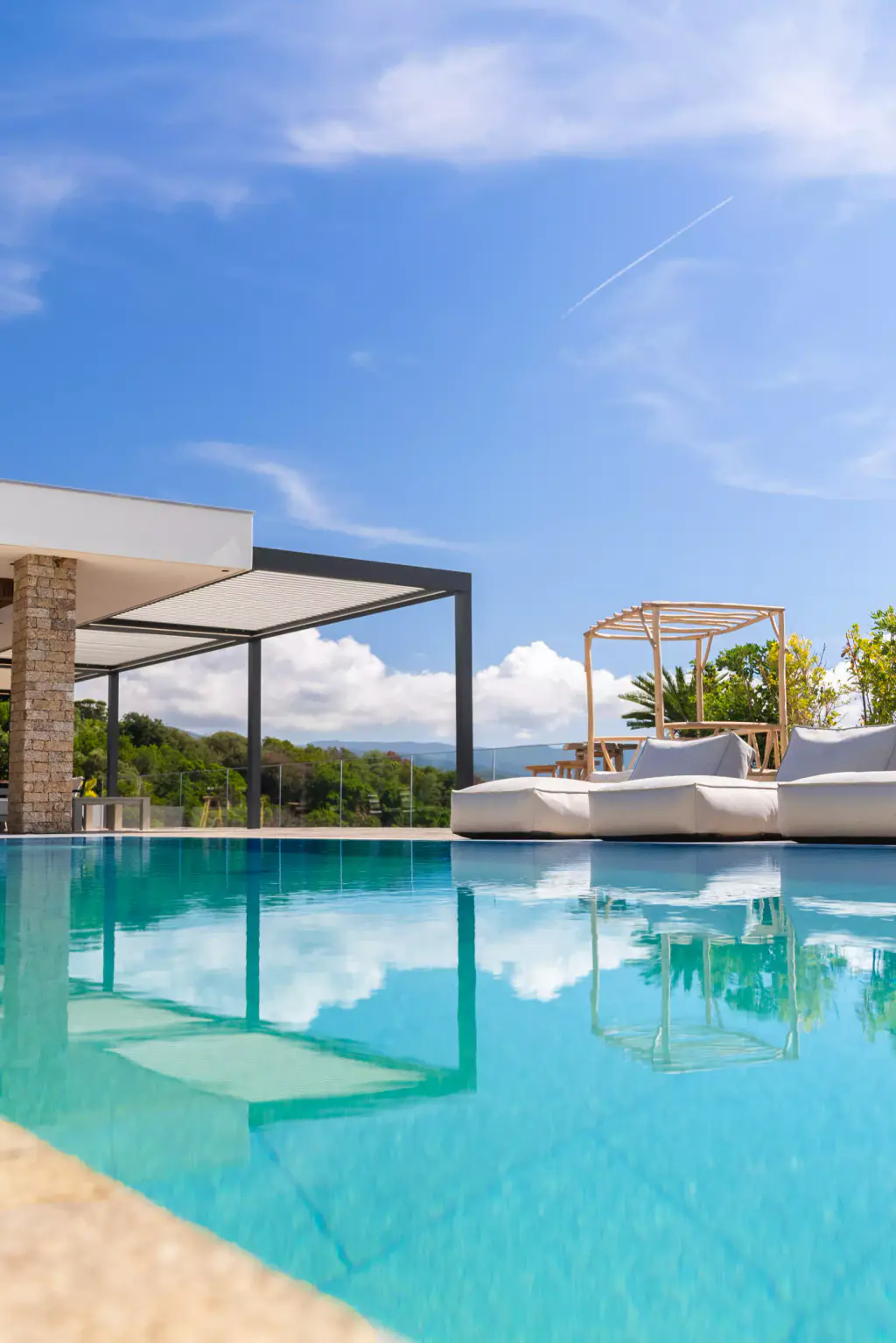 Turquoise pool in the lower half reflecting the clouds, stone coping, bioclimatic pergola, four-poster bed and white sun loungers beyond.