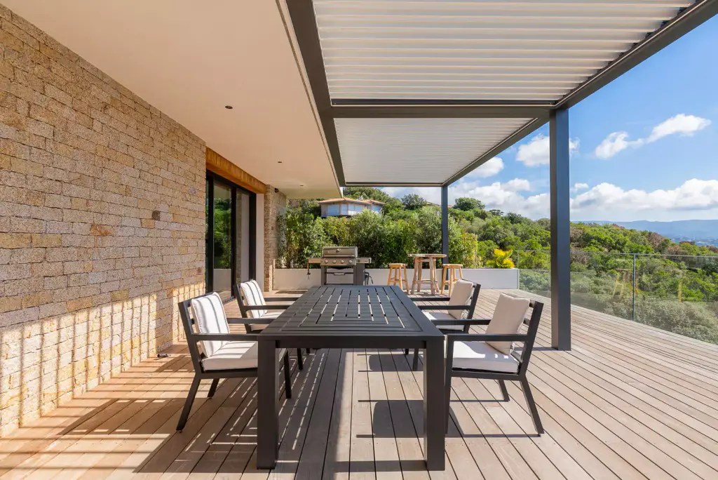 Grande table à manger anthracite et fauteuils sous la pergola bioclimatique, deck en bois, mur de pierre sèche à gauche éclairé par bandes, barbecue et mer au fond.