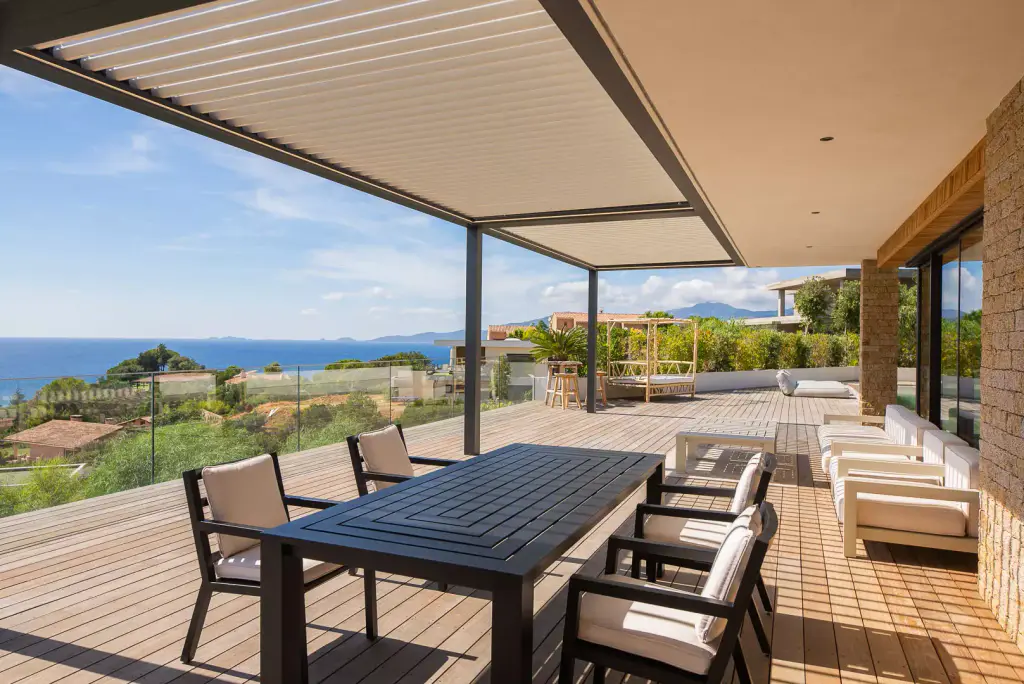 Table à manger anthracite et fauteuils à coussins clairs sous la pergola bioclimatique, deck en bois, salon d’extérieur au fond.
