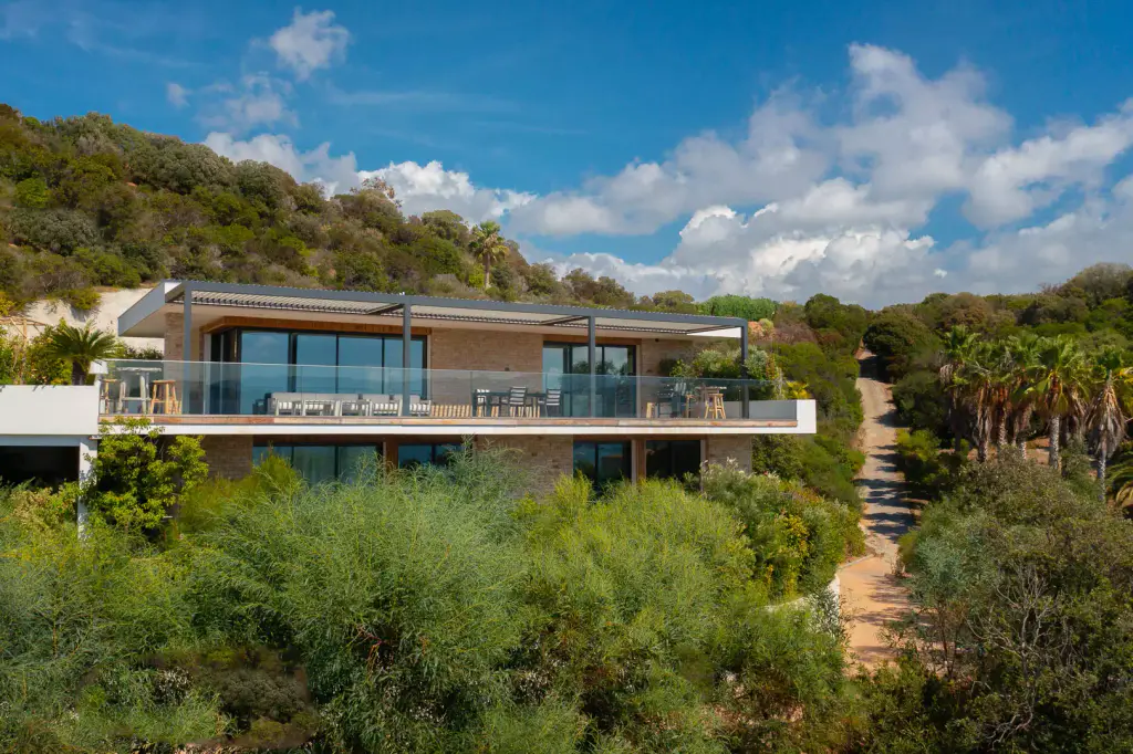 Contemporary villa above the maquis, seen by drone: bioclimatic pergola with an anthracite frame across the whole upper terrace, glass balustrade, wooded hill, cumulus.