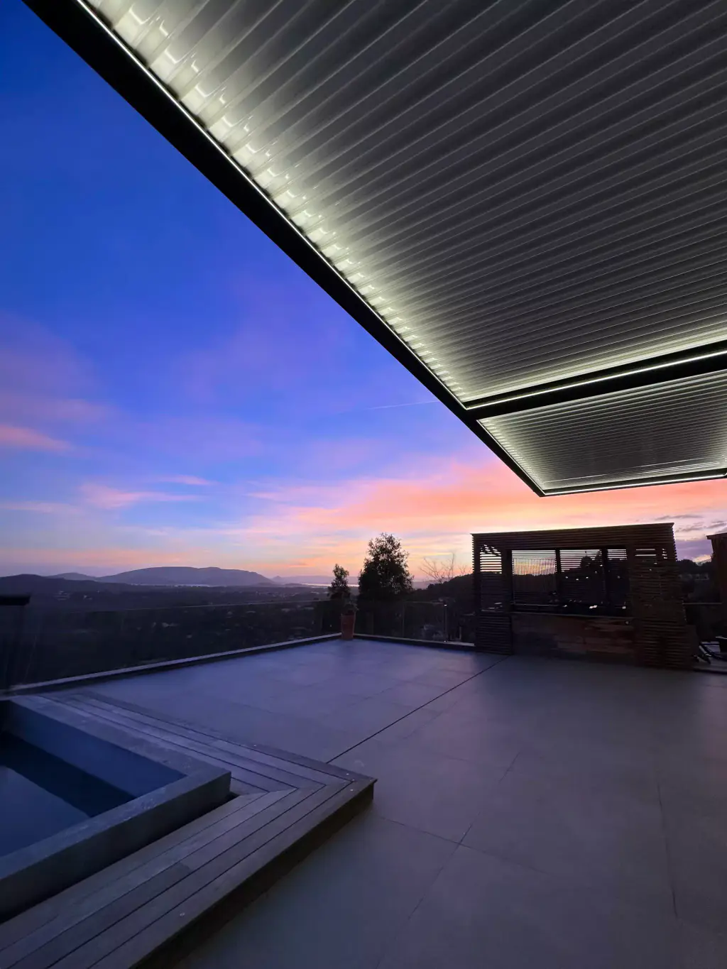 Une pergola dont le ruban lumineux est allumé au crépuscule, au-dessus d’une terrasse, sous un ciel violet.