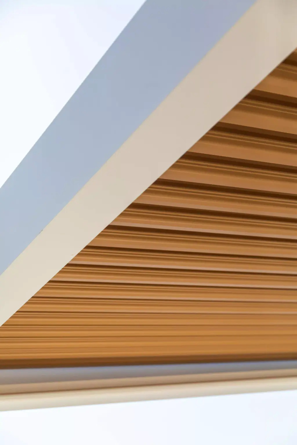 Détail d’angle : cadre blanc et lames effet bois fermées contre un ciel blanc.