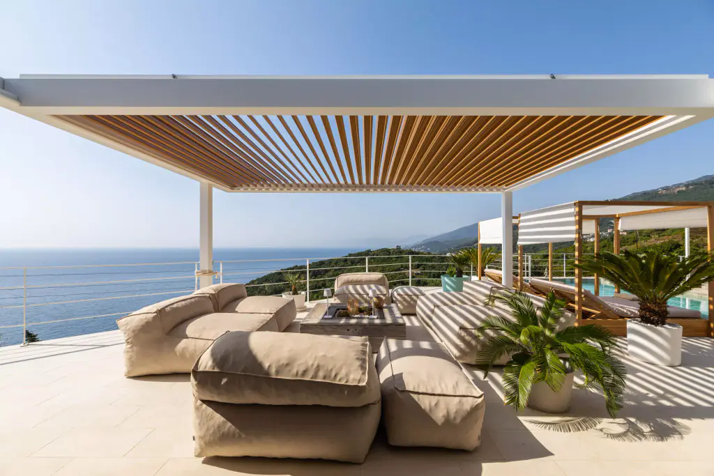 La pergola de la Casa Mare en plein soleil : structure blanche, lames effet bois ouvertes, salon, mer à l’horizon.