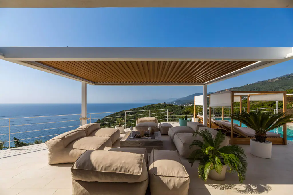 La pergola de la Casa Mare en plein jour, de face : structure blanche, lames fermées, salon de poufs, la mer à l’horizon.