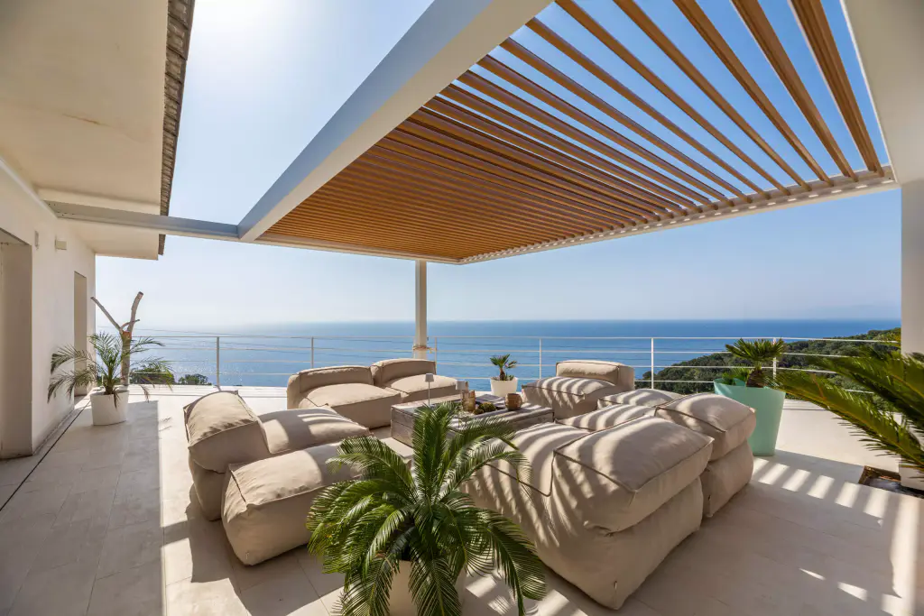 Pergola blanche à lames effet bois ouvertes, vue en contre-plongée, la mer au fond.