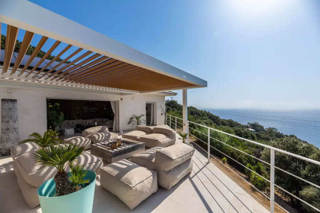 Pergola de trois quarts, lames ouvertes, salon, garde-corps blanc, maquis et mer.