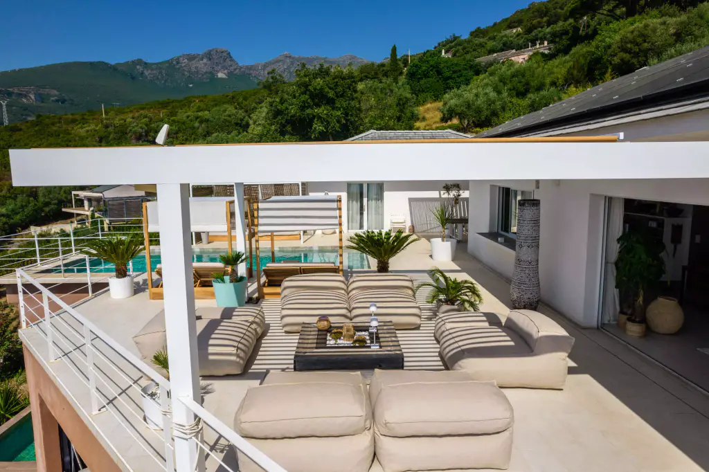 Vue drone basse, de côté sous la pergola : salon, piscine, montagnes.