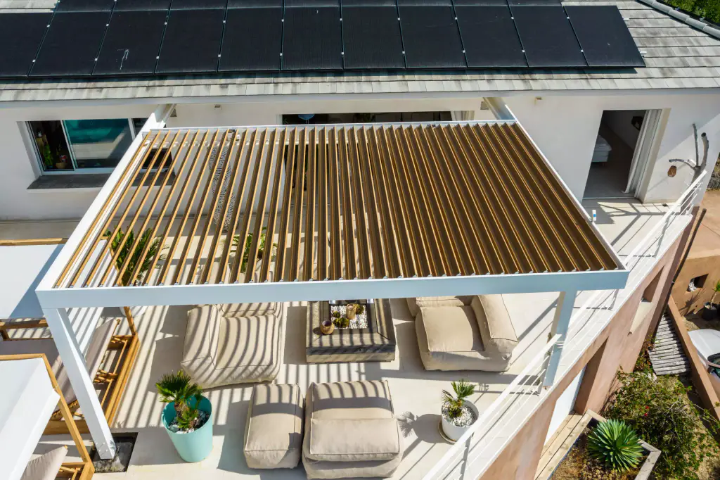 Vue drone de trois quarts : pergola vue du dessus, lames ouvertes laissant voir le salon, panneaux solaires de la villa.