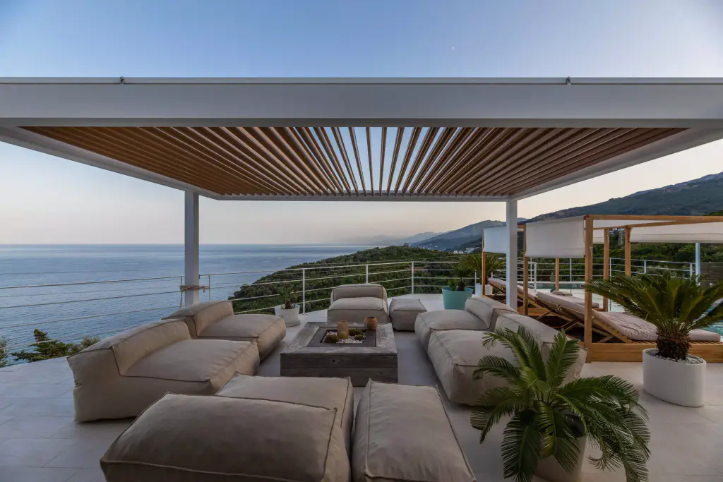 La pergola de la Casa Mare au crépuscule : lames ouvertes sur un ciel rosé, salon d’extérieur, mer calme.
