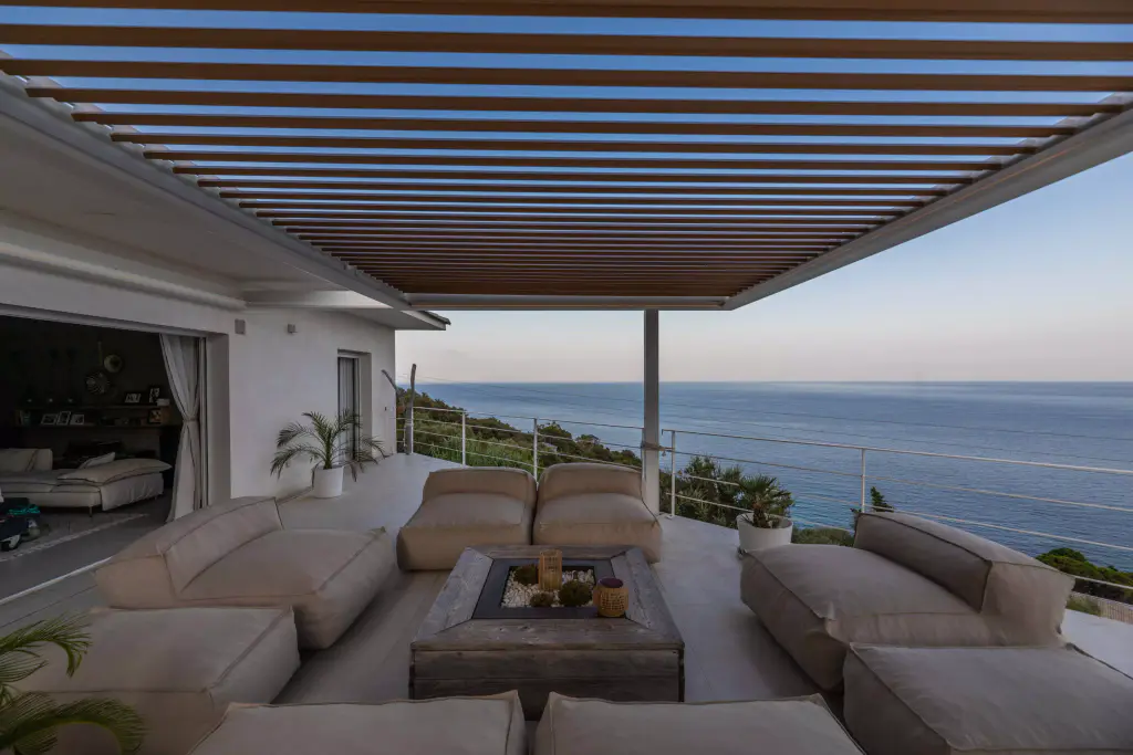 Vue frontale sous la pergola au crépuscule, lames ouvertes, salon et mer.
