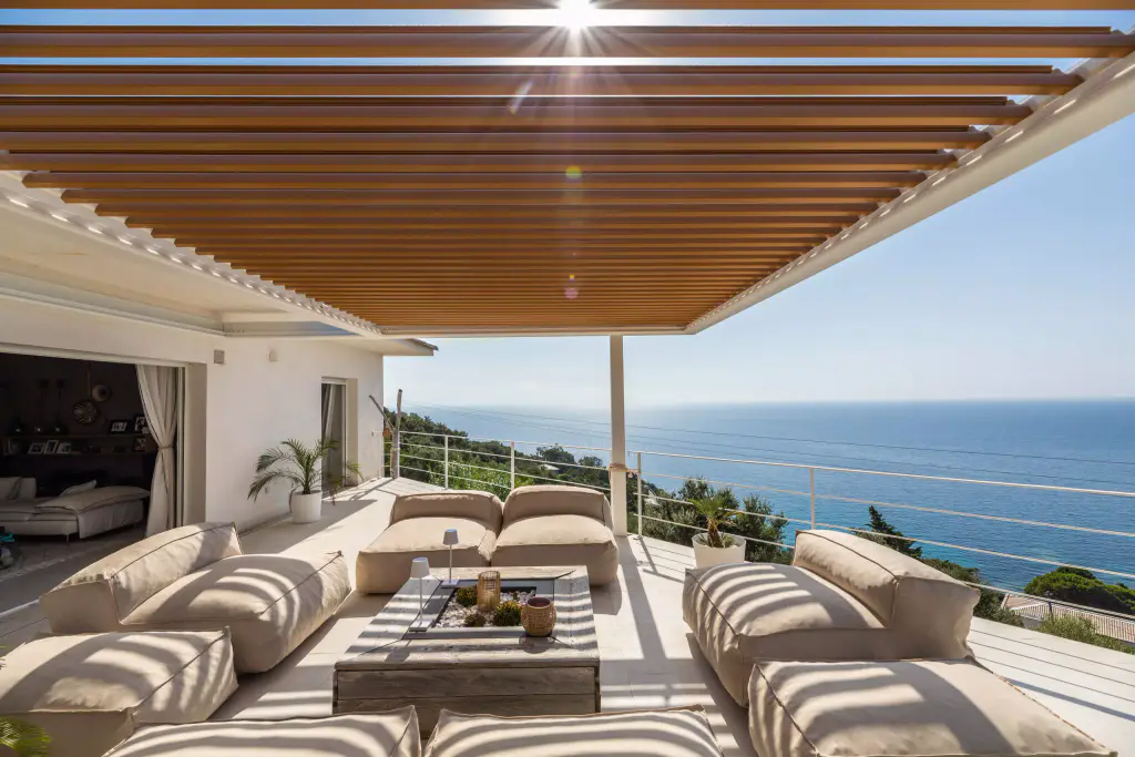 Vue frontale sous la pergola : une étoile de soleil perce entre les lames, salon, mer.