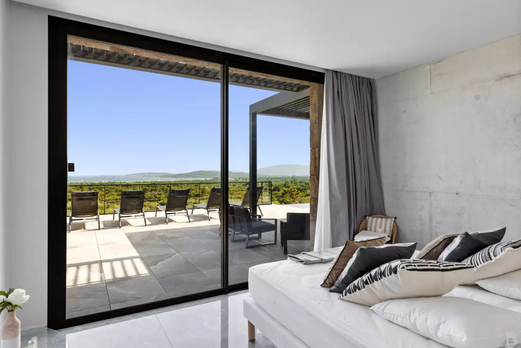 Bedroom: bed and cushions, large black sliding glazing opening onto the terrace and its loungers, grey sheer curtain, concrete wall.