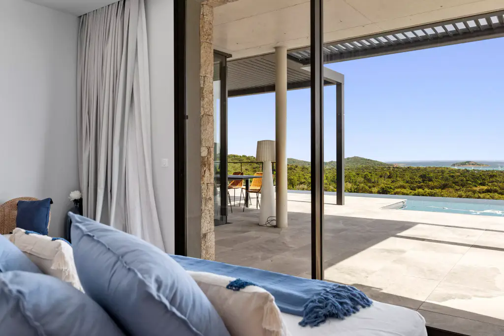 Bedroom: blue cushions in the foreground, glazing open onto the terrace, pergola visible at top right, pool.