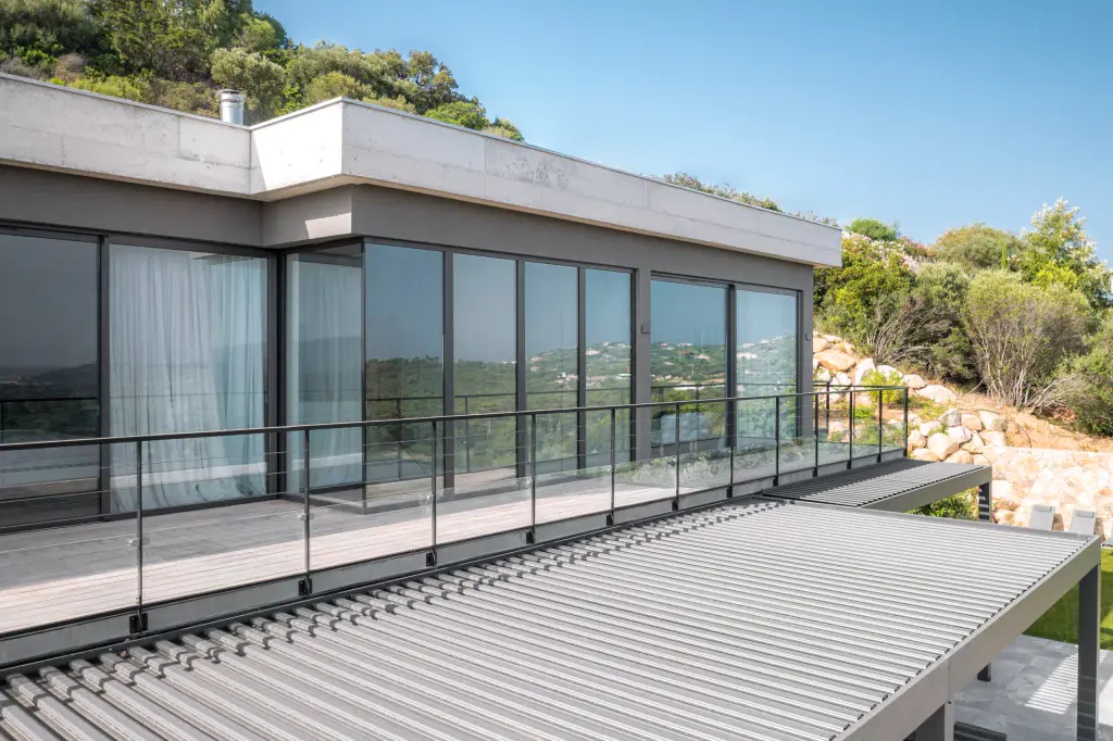 Three-quarter drone view: pergola louvres in the foreground, glazed upper floor, maquis.