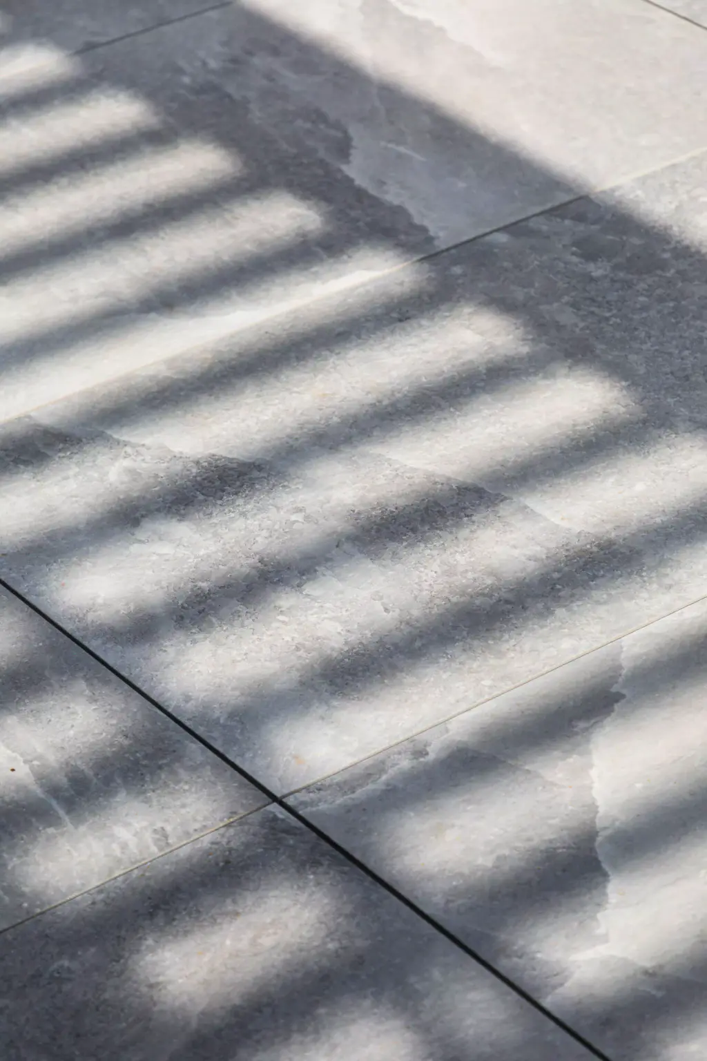 Ombre rayée des lames de la pergola sur le dallage de grès gris de la terrasse, en gros plan, un joint de dalle traversant l’image en diagonale.