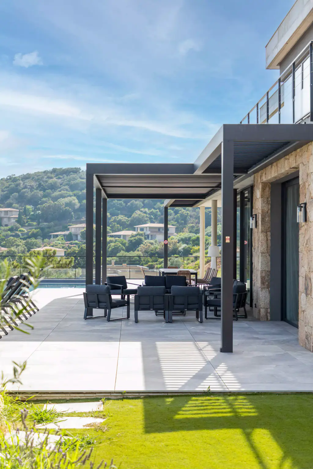 Pergola anthracite adossée à la villa en pierre, salon d’extérieur dessous, pelouse au premier plan avec l’ombre des lames.