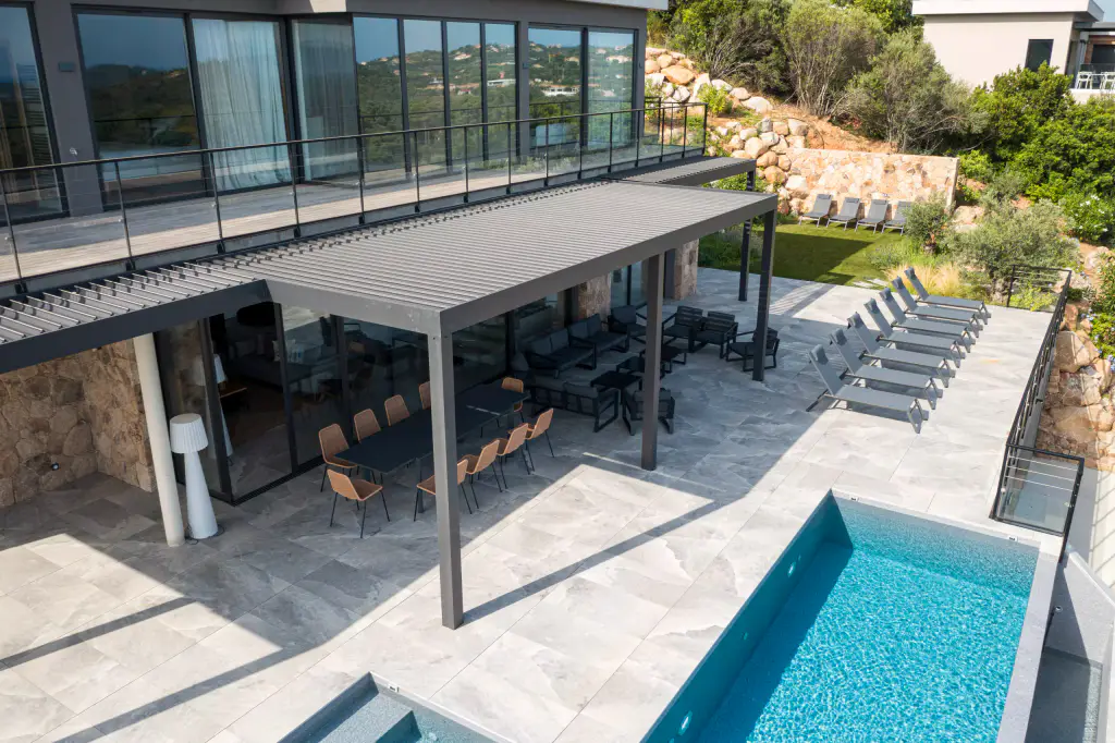 Vue drone plongeante : pergola anthracite, table à manger dessous, transats, piscine turquoise, dallage gris clair.