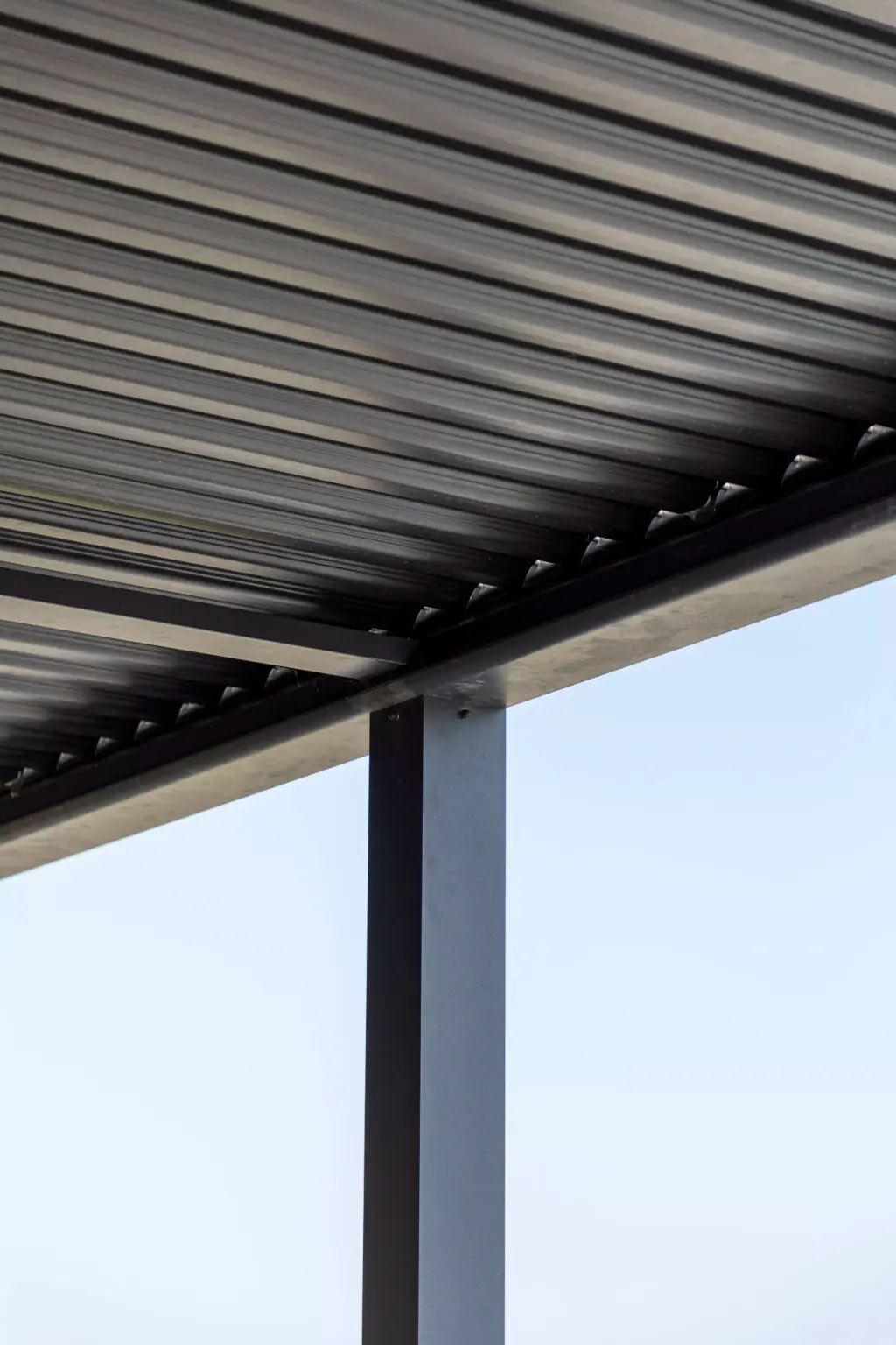 Anthracite bioclimatic pergola louvres, closed, seen from a low angle, with the post and the frame beam outlined against a very bright sky.