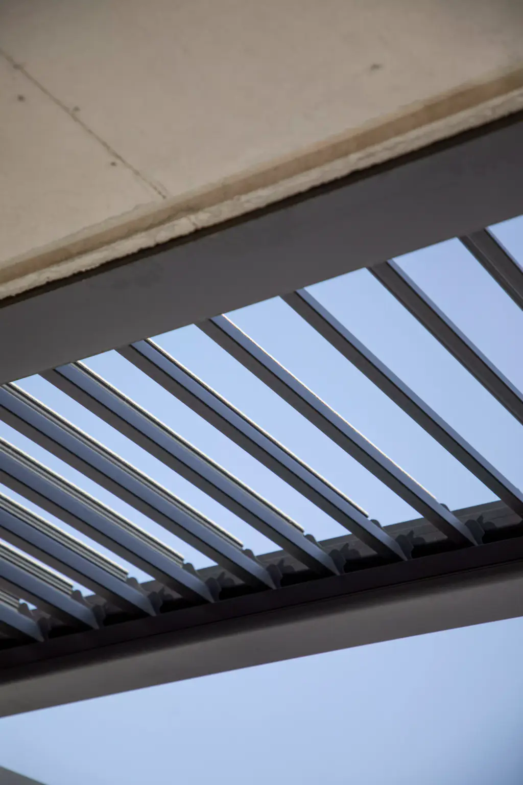 Anthracite bioclimatic pergola louvres, open, seen from below against a pale sky, between the concrete underside of the house and the frame beam.