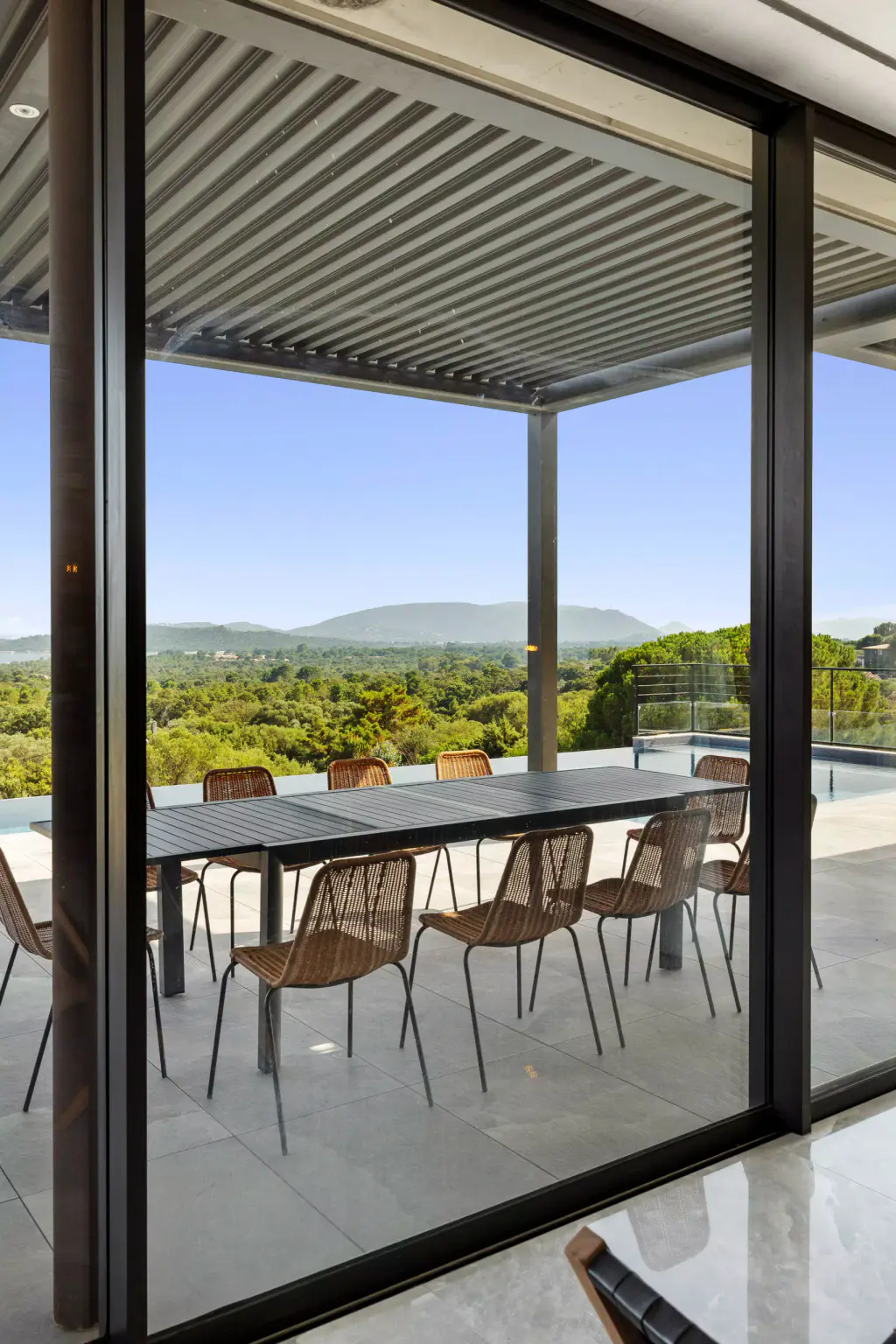 Under the pergola seen from inside: large table and rattan chairs, maquis and the distant sea.