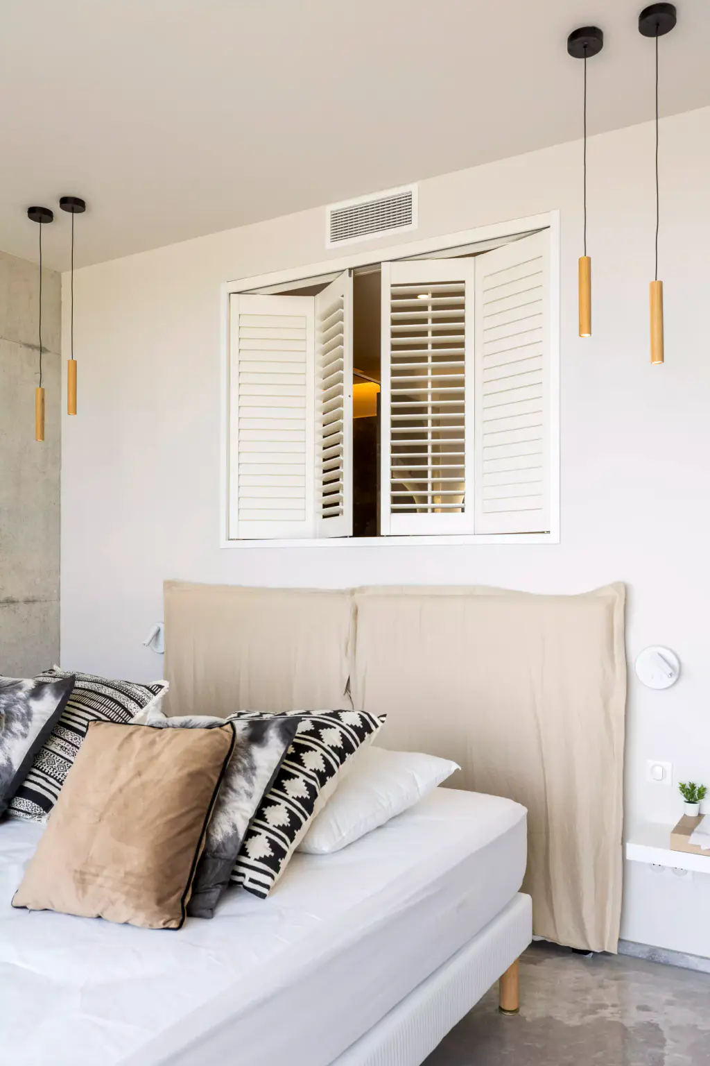 Persiennes intérieures blanches entrouvertes au-dessus d’une tête de lit beige, entre deux suspensions en bois ; coussins à motifs noirs et blancs sur le lit, murs blancs.