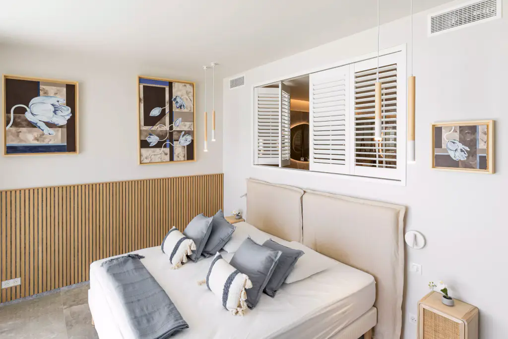Persiennes intérieures blanches à lames orientables au-dessus d’un lit, dans une chambre claire : tête de lit beige, soubassement en tasseaux de bois, tableaux encadrés.