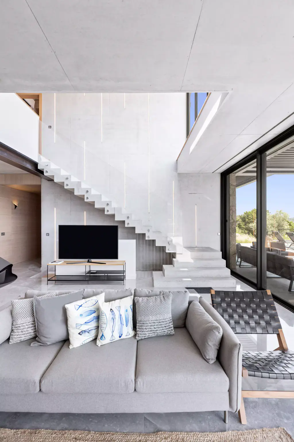 Double-height concrete living room: floating staircase, grey sofa, television.