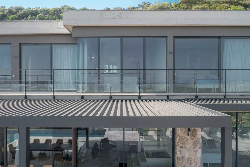 Front drone view: pergola roof closed in the foreground, glazed upper-floor façade and balustrade.