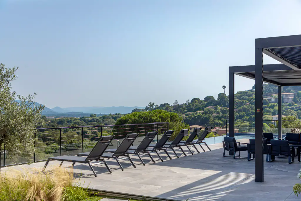 Terrasse : alignement de transats, pergola anthracite abritant un salon, collines sous un ciel voilé.