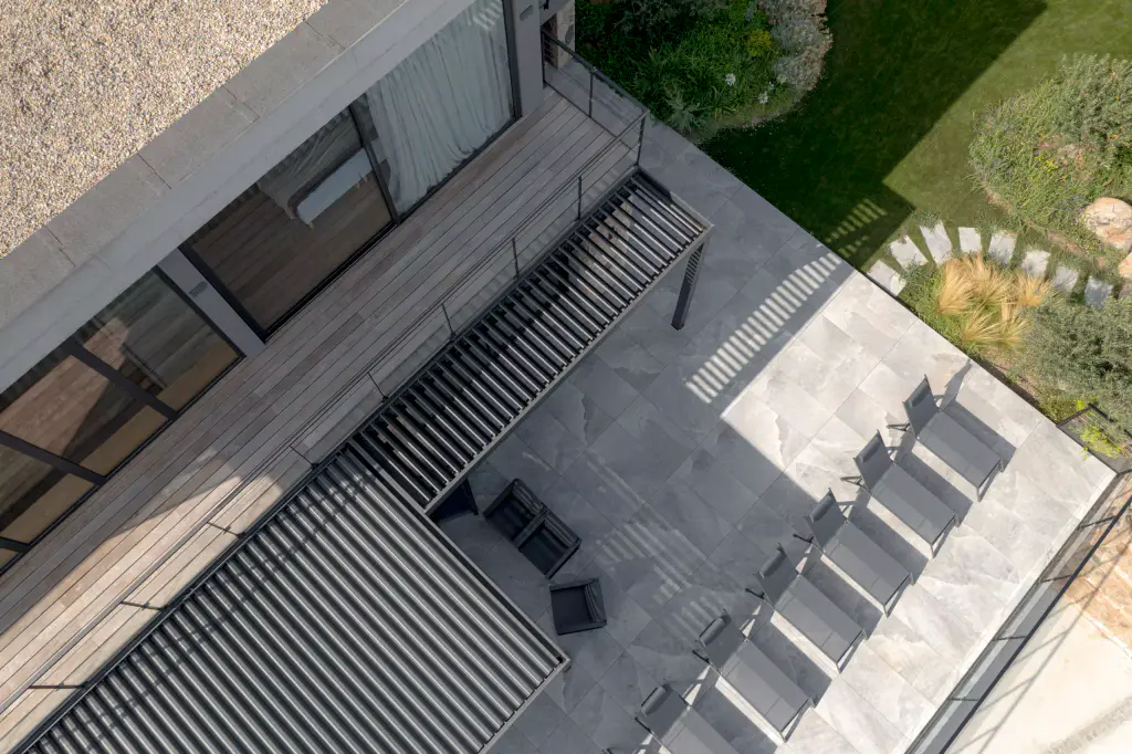 From the air, on the diagonal: the anthracite louvres of the pergola, the row of sun loungers, the grey paving, the lawn.