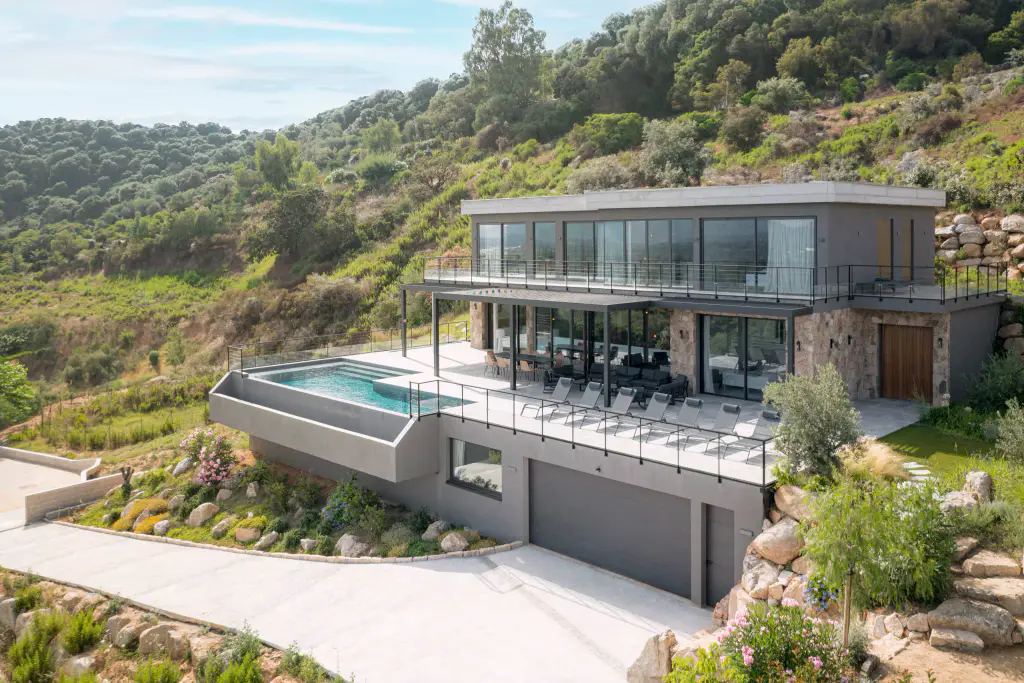 Vue drone large : la villa posée dans la colline, piscine à débordement, garage, jardin fleuri.