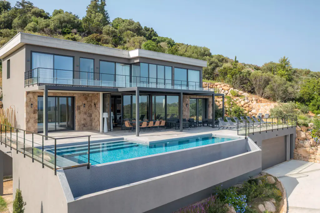 Vue drone de trois quarts : villa contemporaine sur deux niveaux, piscine à débordement, pergola anthracite devant le rez-de-chaussée, maquis en fond.