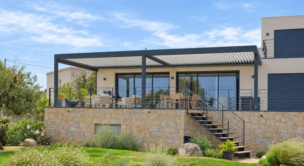 Pergola bioclimatique gris anthracite adossée à une villa contemporaine, sur une terrasse haute à soubassement de pierre, avec garde-corps à lisses et escalier vers le jardin.