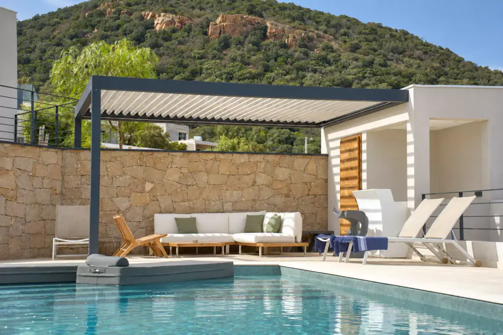 Pergola bioclimatique gris anthracite au bord d’une piscine, lames claires entrouvertes au-dessus d’un canapé d’angle blanc, mur de pierre et colline rocheuse boisée derrière.