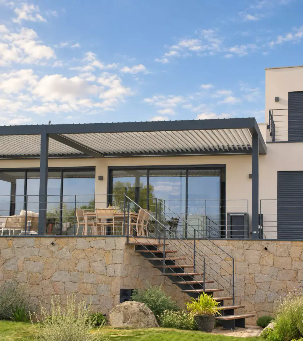 Pergola bioclimatique gris anthracite sur la terrasse haute d’une villa contemporaine, baies vitrées derrière, soubassement de pierre et escalier métallique vers le jardin.